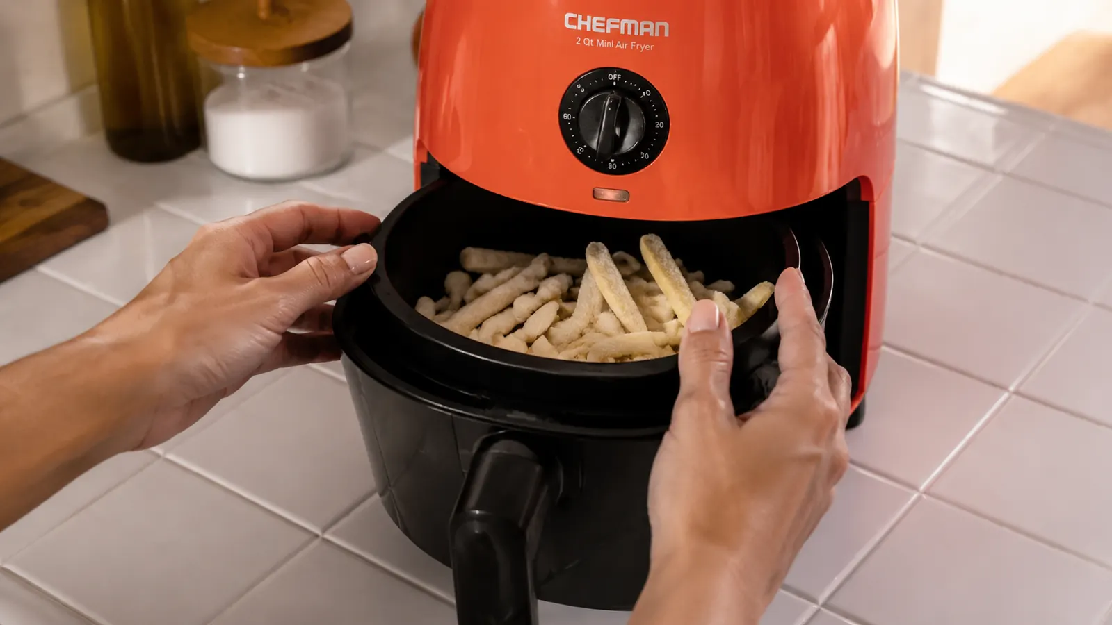 Hands placing a small basket of frozen fries into a compact CHEFMAN air fryer on a white tile counter