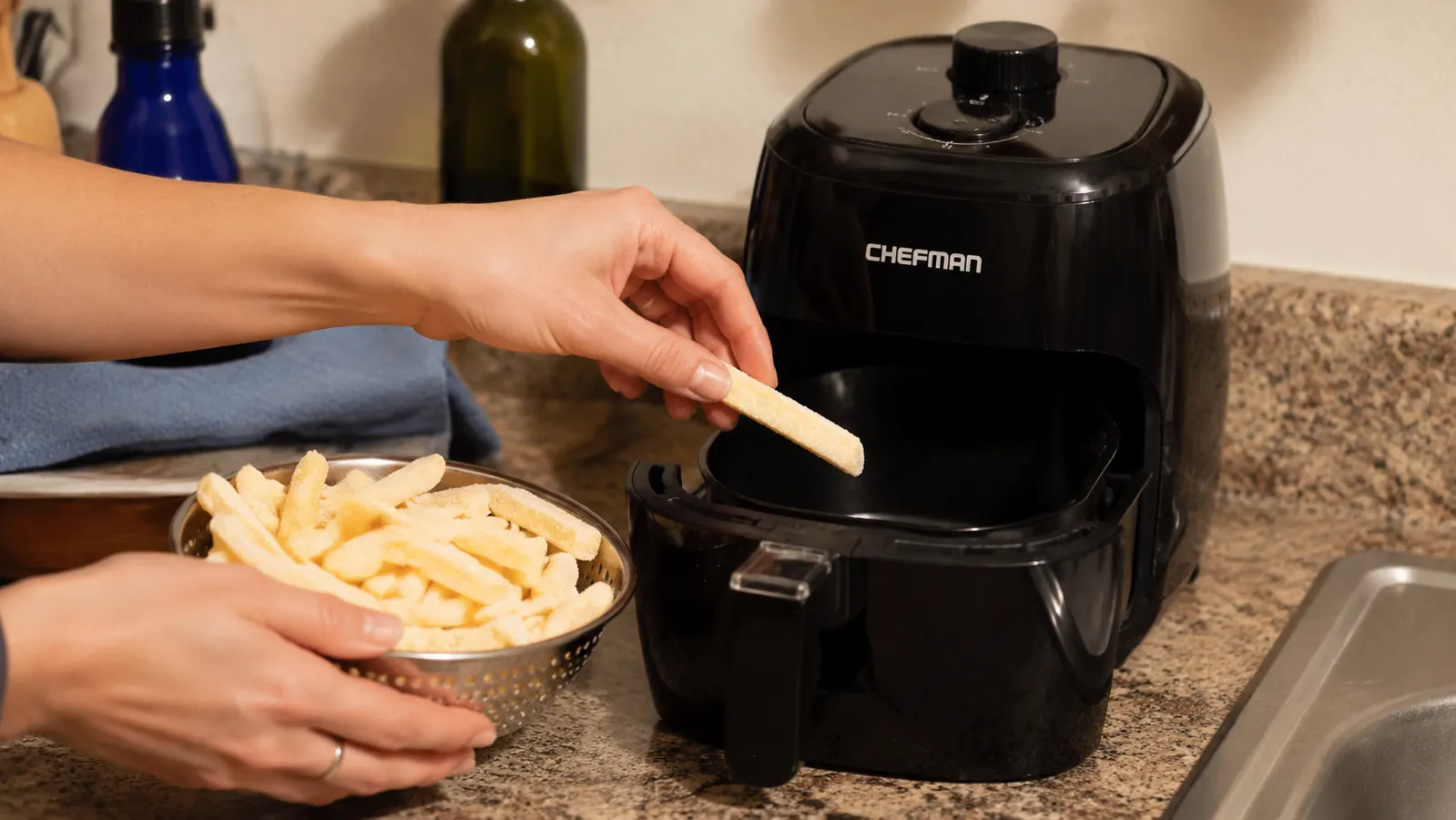 Hand placing a handful of frozen French fries into the CHEFMAN mini air fryer basket over a kitchen counter