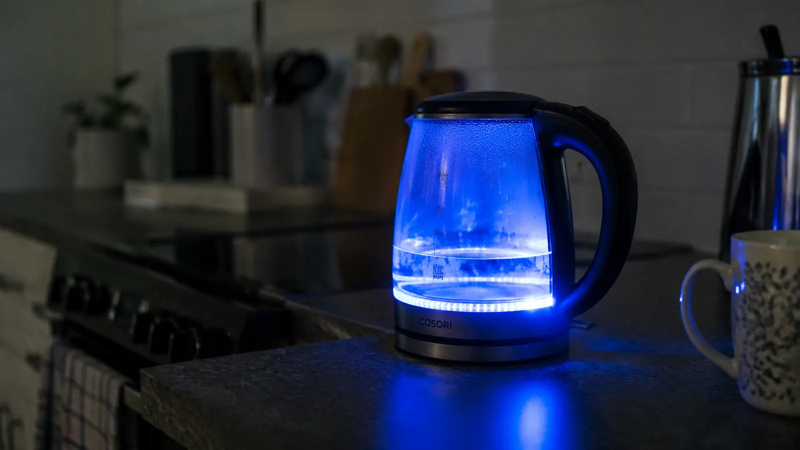 White electric kettle with bright blue LED glow visible through the water window in a dim kitchen